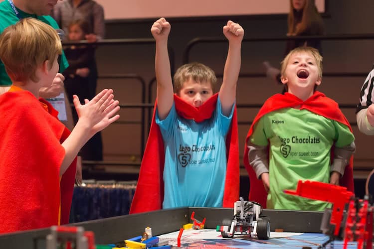23562944380_a8d3063e7c_o-e1723073858852 From right to left, three kids smiling, pumping two fists in the air, clapping at the 2015 Juneau Robot Jamboree - Trash Tech